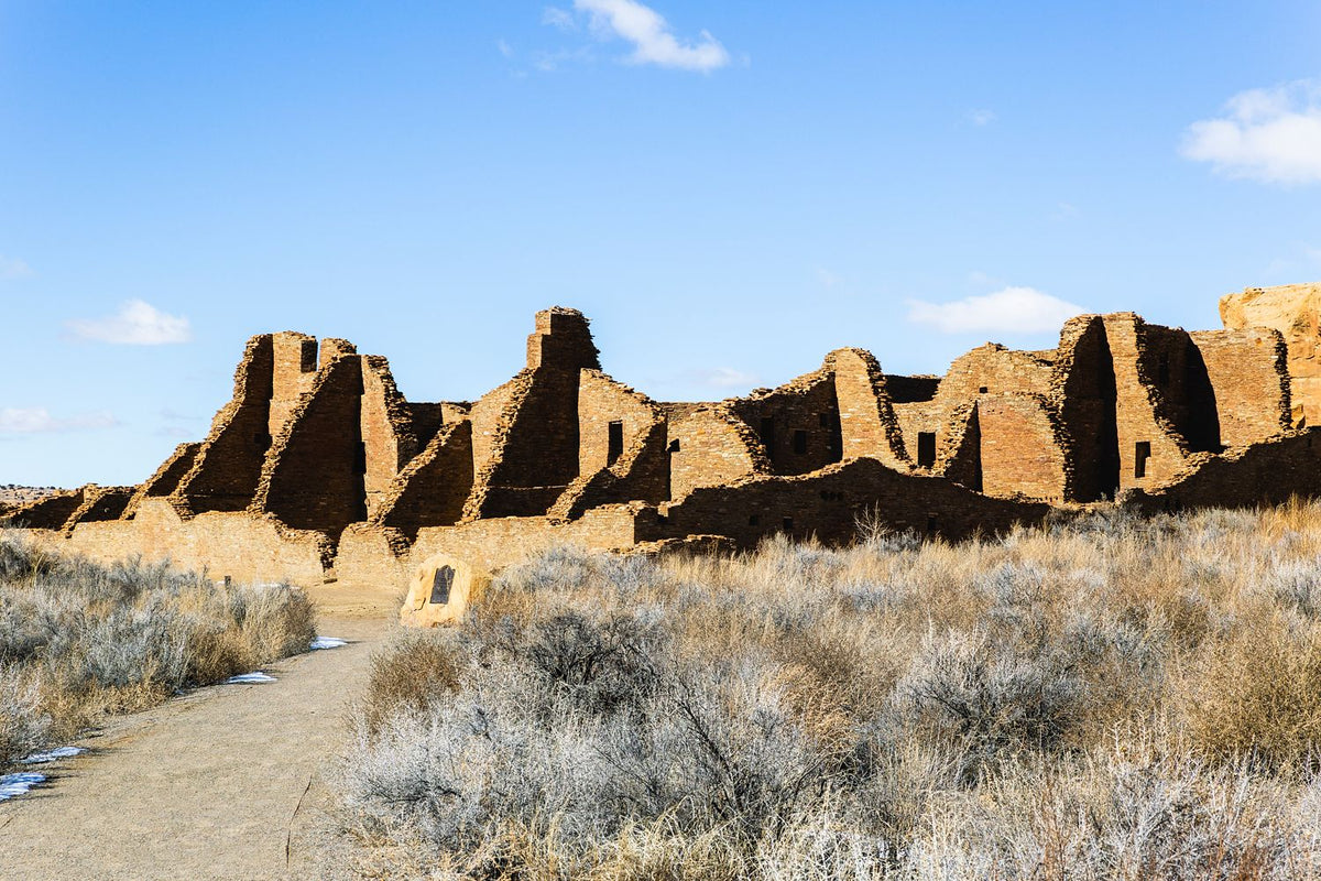 Ancient masonry ruins across a grassy desert landscape beneath a blue sky in Chaco Canyon, New Mexico.