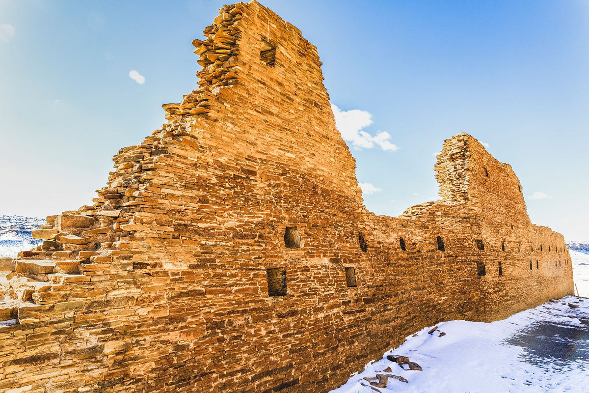 Sunlit stone walls with window openings and snow at their base in Chaco Canyon, New Mexico.