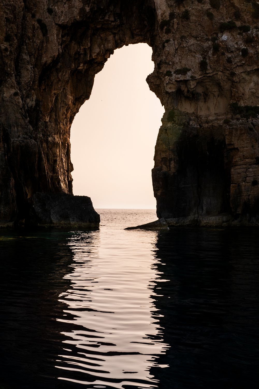 Natural sea arch near Pylos, Greece, with warm light shining through the rock opening and reflecting on calm water.