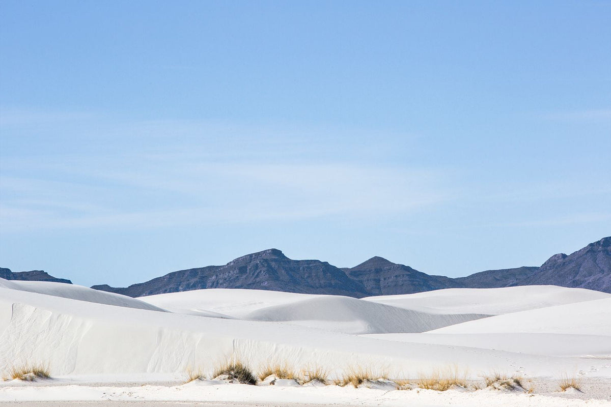 White sand dunes with sparse grasses and distant dark mountains beneath a pale blue sky in New Mexico.