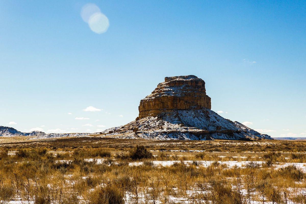 A flat-topped desert butte above golden grasses and patches of snow beneath a blue sky.
