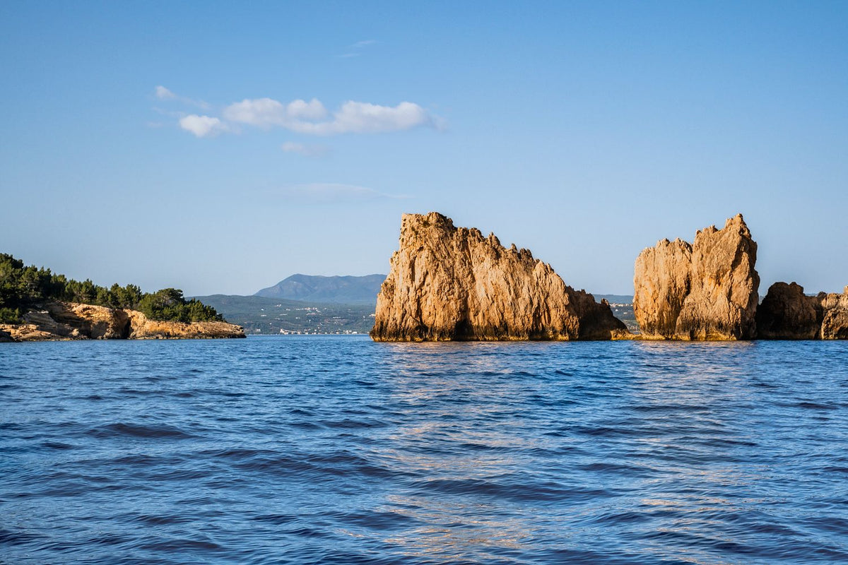 Sunlit sea stacks rising from blue water in Navarino Bay, Messinia, Greece, with a distant coastline beneath a clear sky.
